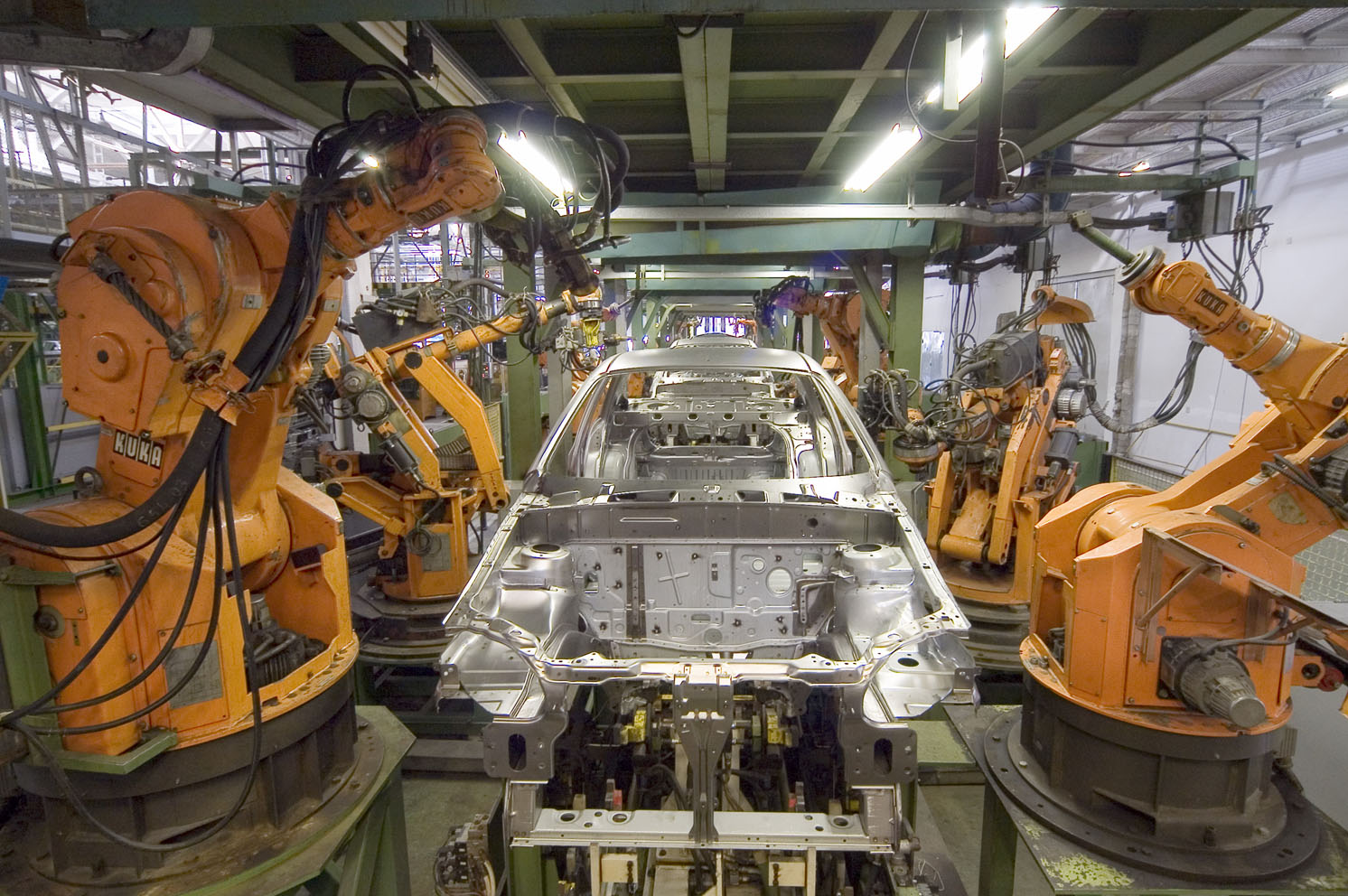 Orange KUKA industrial robot arms on both sides of a bare steel car body on a production line